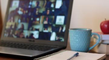 a laptop screen showing an online meeting and a notepad and pen on a desk
